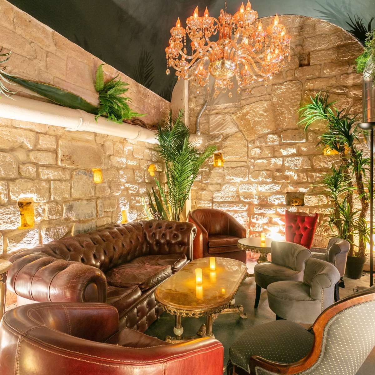 A cozy lounge area with a plush leather sofa, elegant chairs, and decorative plants against a backdrop of stone walls and a chandelier.