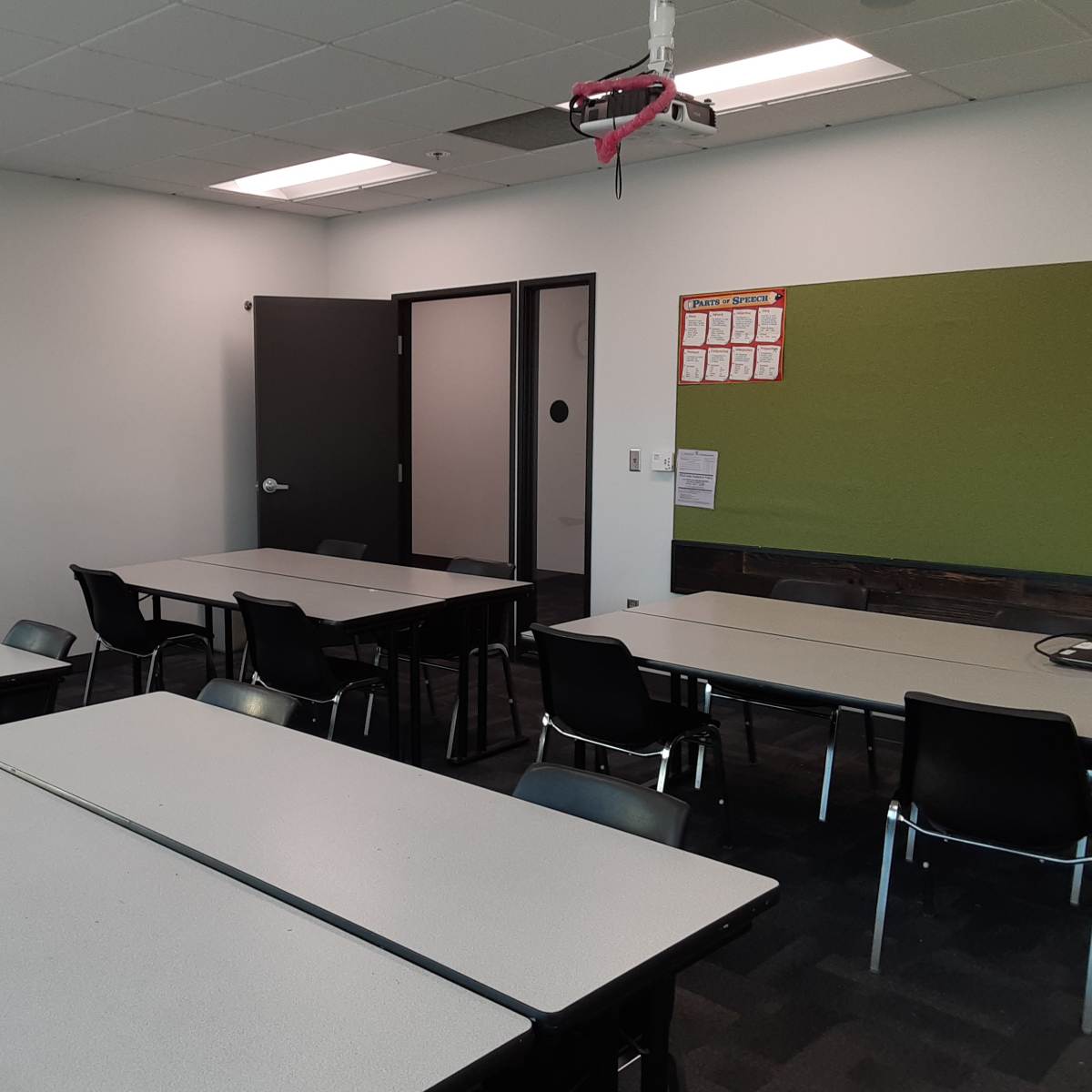 A classroom features several tables arranged with chairs, a clock on the wall, a green board, and a door leading to another room.