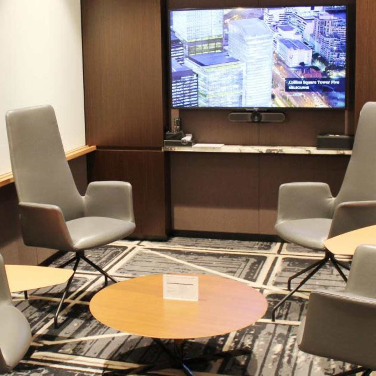A modern seating area with four gray high-back chairs arranged around a circular wooden table, accompanied by a large screen displaying a building image on the wall.