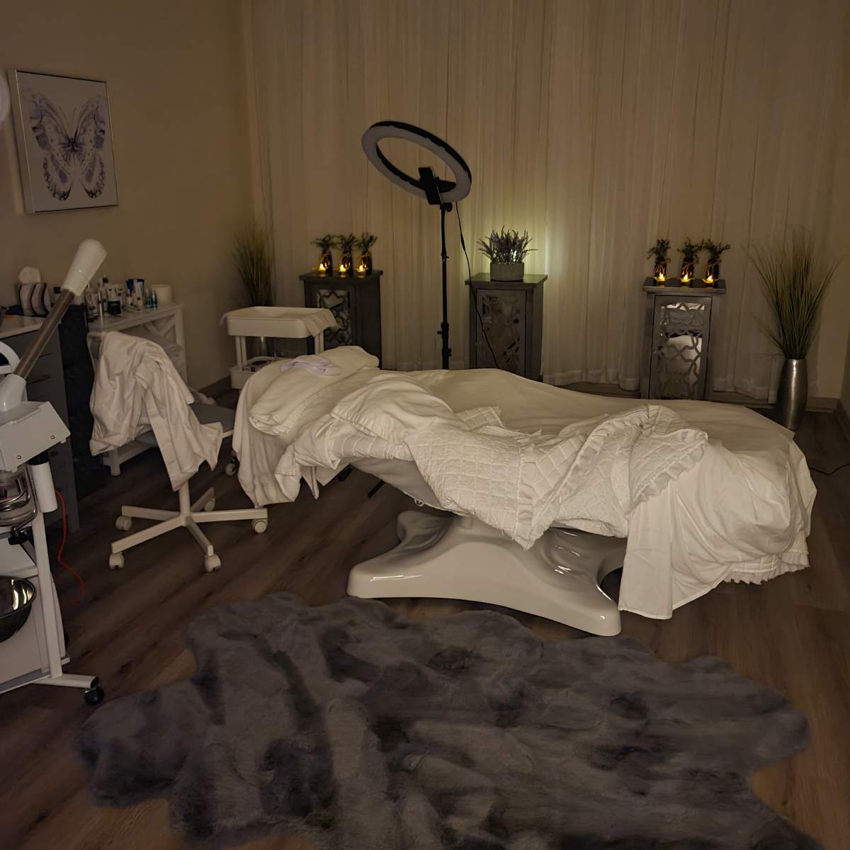 A spa treatment room with a massage table covered in white linens, surrounded by a stool, a floor rug, and softly lit decor elements.