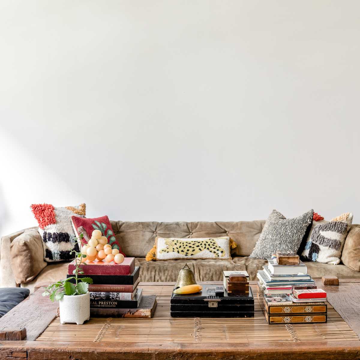 A spacious living room features a large sofa adorned with decorative pillows, a wooden coffee table stacked with books and a plant, and a wall painted in a neutral color.