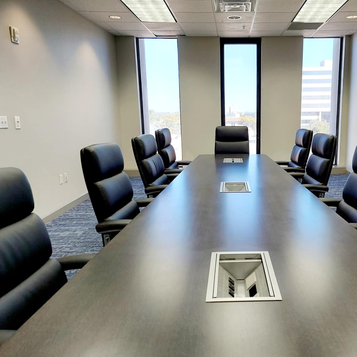 A modern conference room features a long dark table surrounded by eight leather chairs, with large windows providing a view of the cityscape.