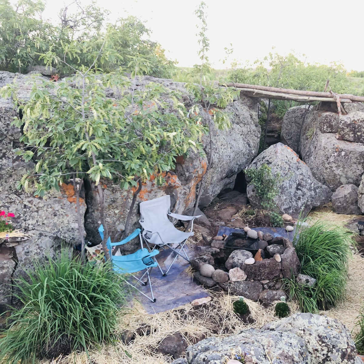 The scene features a small outdoor seating area with two folding chairs near a circular fire pit surrounded by large rocks and greenery.