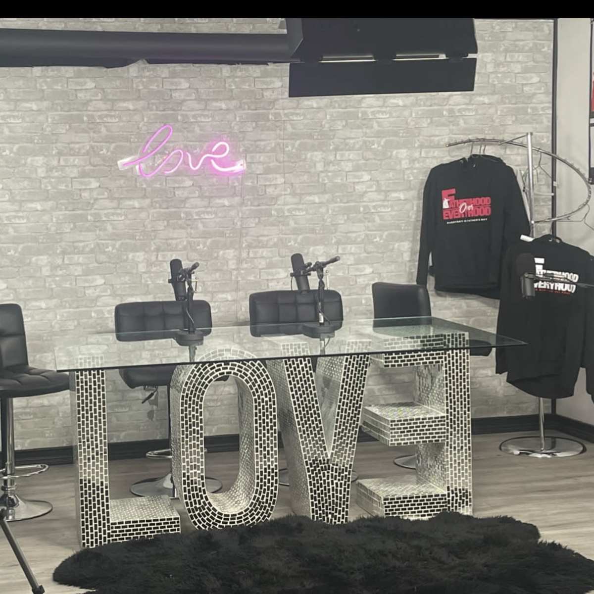 A modern podcast studio features a glass table with the word "LOVE" made from mirrored tiles, two microphones, and a neon sign above the table.