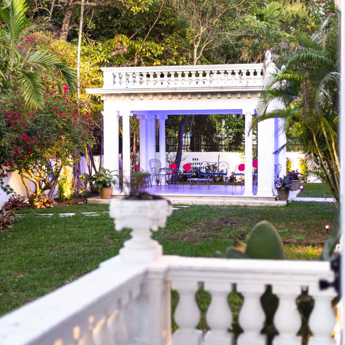 A white gazebo with a decorative railing and seating area is surrounded by lush greenery and colorful flowering plants.