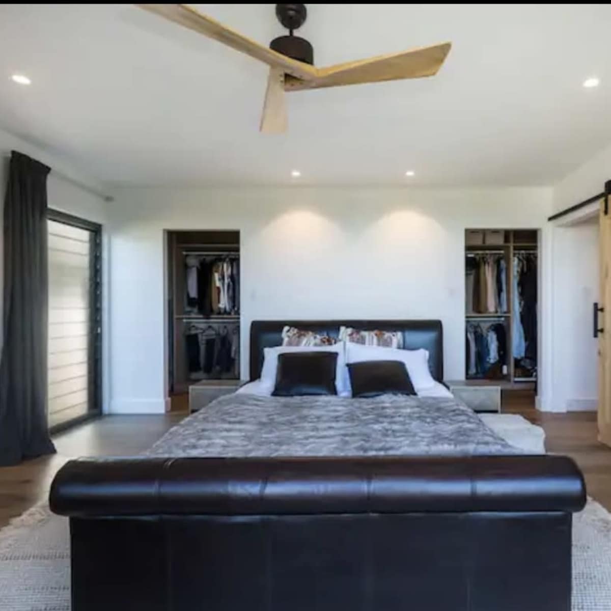 The bedroom features a large bed with a dark leather bench at the foot, a ceiling fan, and a sliding wooden door leading to a closet.