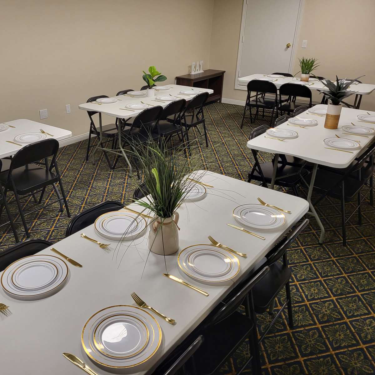 A spacious room is set up with multiple tables, each arranged with white plates, gold utensils, and potted plants as centerpieces.