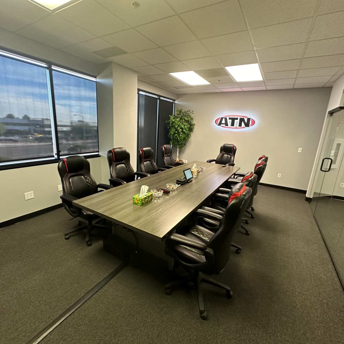 A modern conference room features a large table surrounded by black executive chairs and large windows providing natural light.