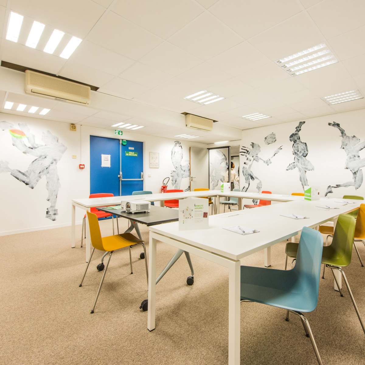 A modern meeting room with a long table surrounded by colorful chairs, and walls decorated with black and white abstract figures.