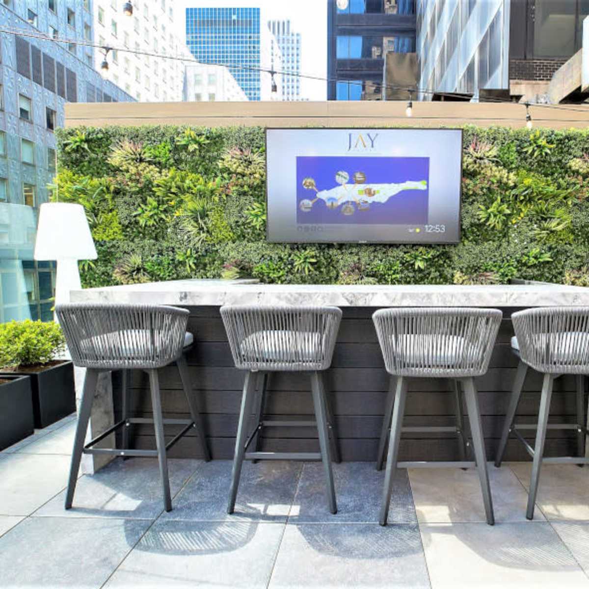 A modern rooftop bar area with a stone counter and four high chairs, surrounded by green wall planters and featuring a large flat-screen TV.