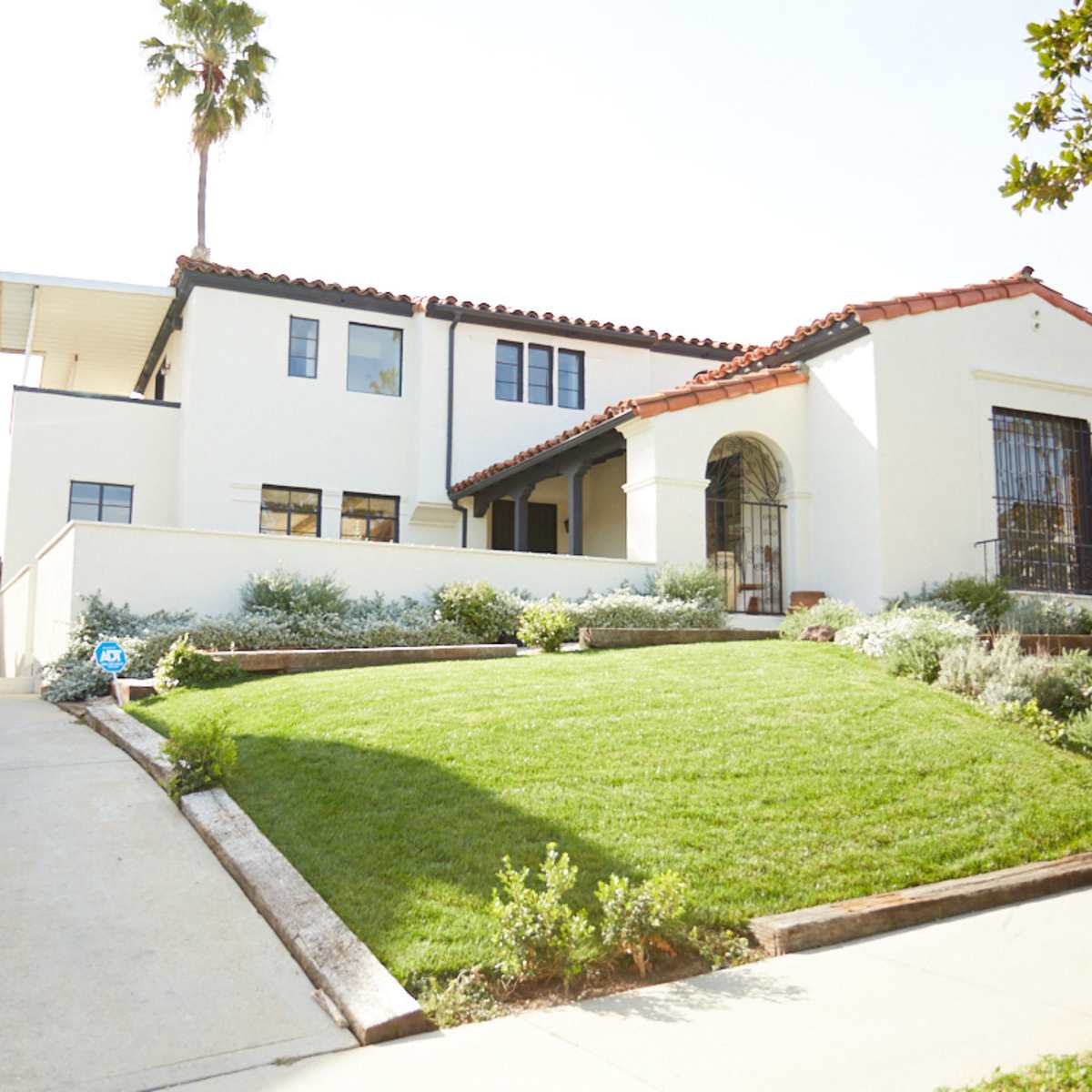 A white house with a red tile roof and a decorative archway sits on a landscaped lot with a sloped driveway and palm trees nearby.
