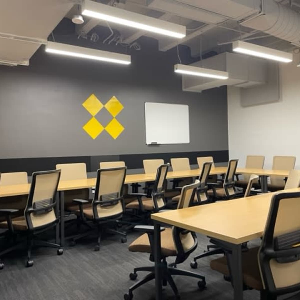 A conference room features multiple wooden tables and chairs arranged in rows, with a whiteboard and a yellow diamond graphic on the wall.