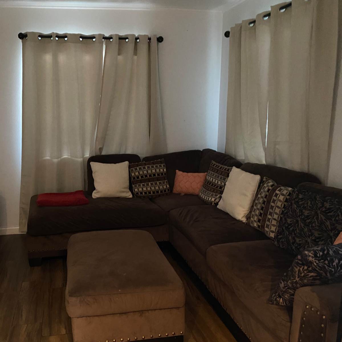A sectional sofa with various decorative pillows is positioned in a living room with light-colored walls and sheer curtains, alongside a television and a small storage basket.