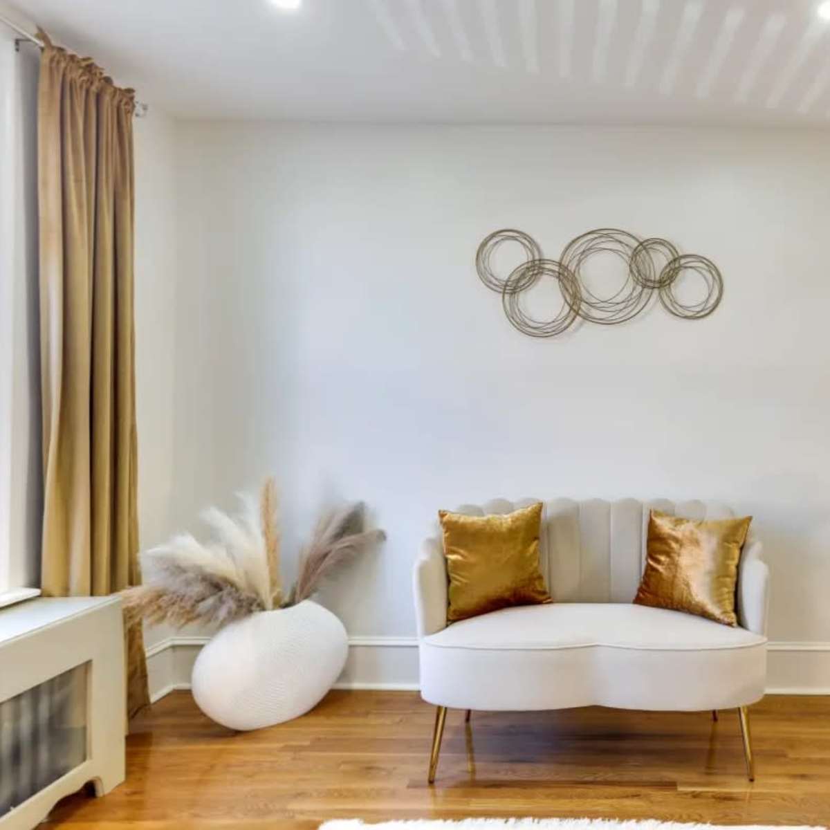 A minimalist living room setup featuring a light-colored loveseat with decorative cushions, a textured wall hanging, and a decorative vase beside a staircase.