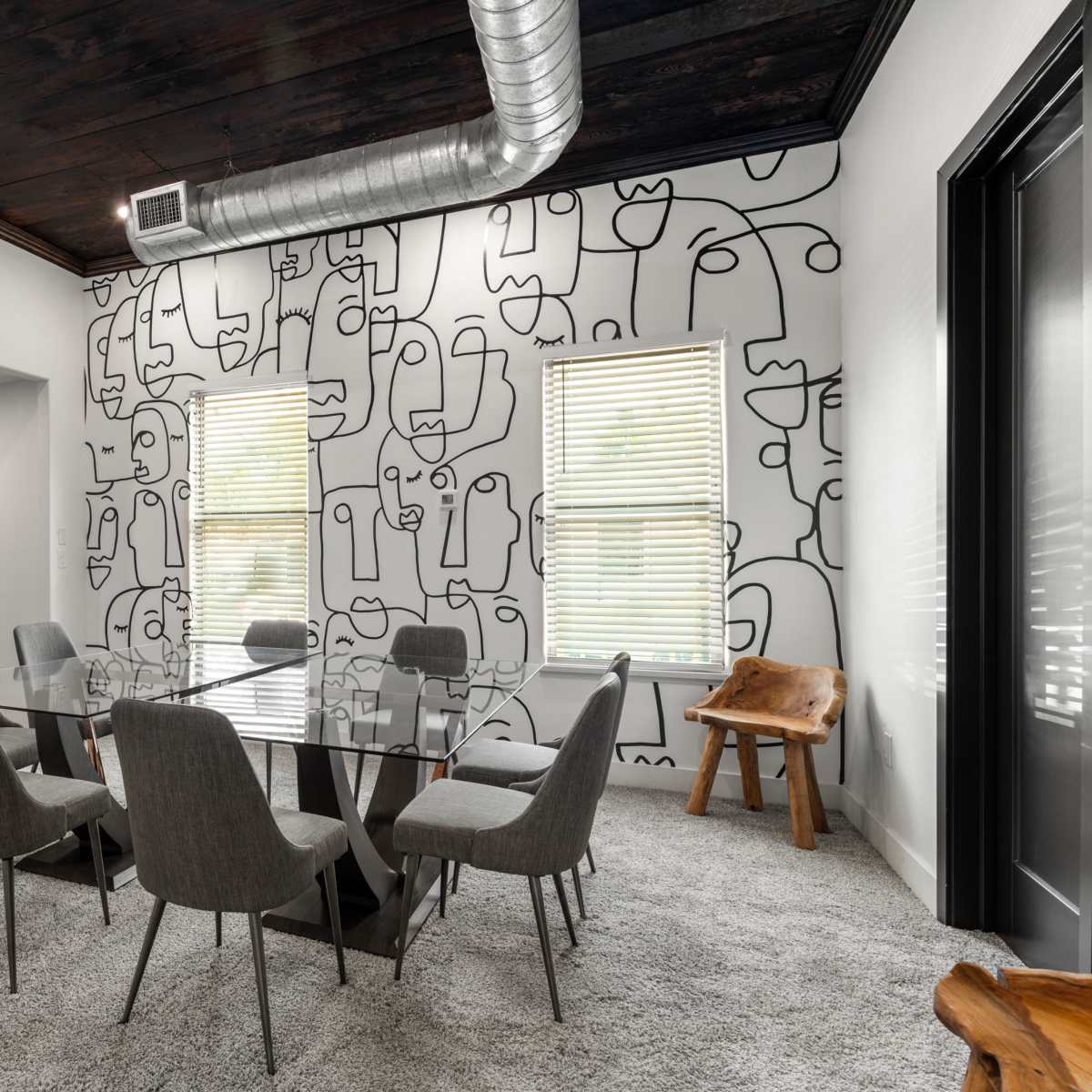 A dining area features a glass table surrounded by six chairs, with a wall adorned by a minimalist line drawing design and a wooden accent piece in the corner.