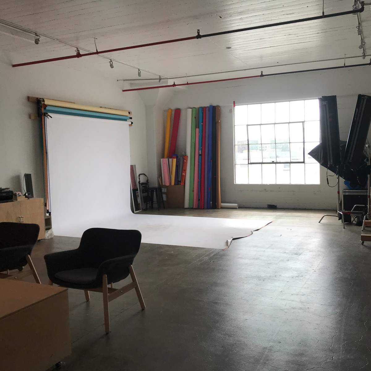 A photography studio with a white backdrop, colorful paper rolls, a large window, and several furniture items.
