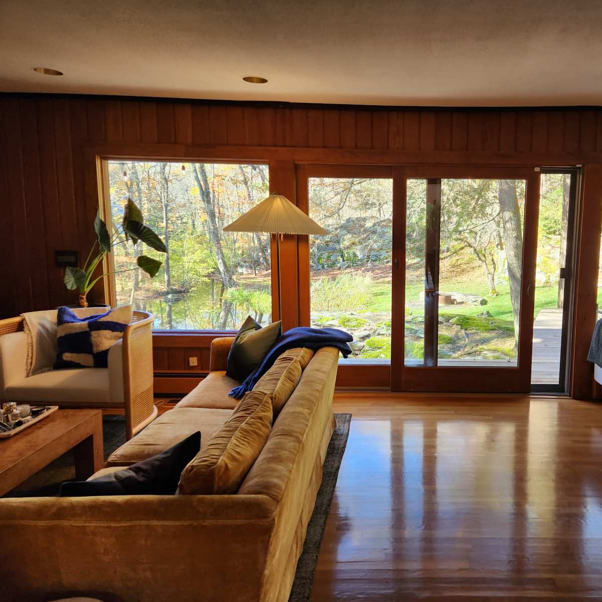A spacious living room features large windows overlooking a natural landscape, with a mix of comfortable seating and decorative elements.