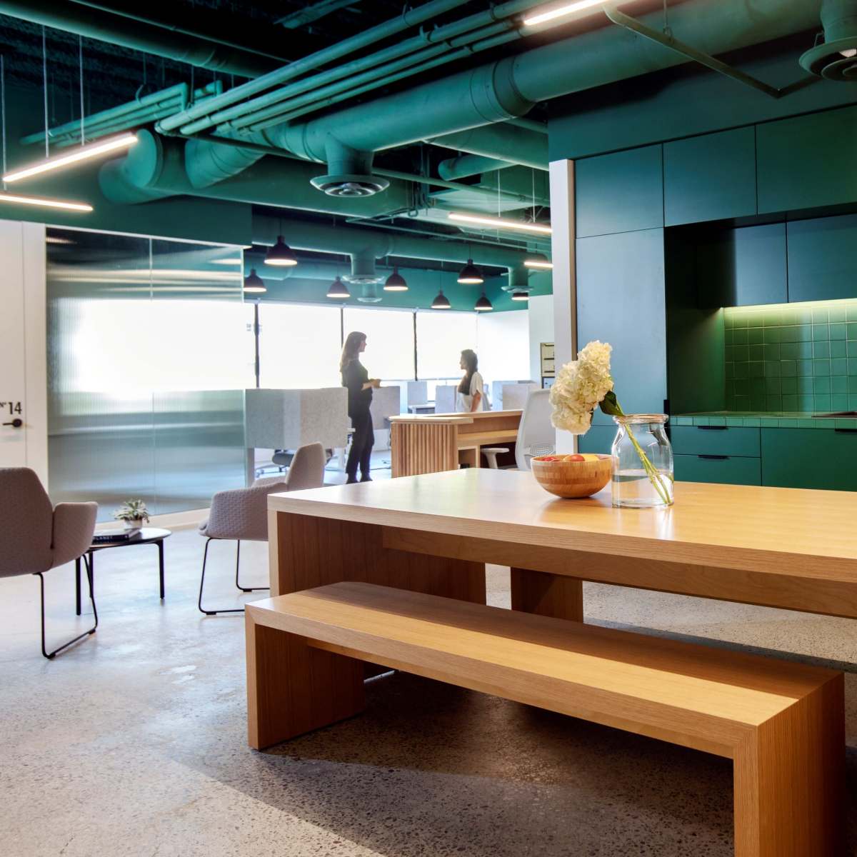 A modern office kitchen features a large wooden table, green cabinetry, and a tiled backsplash, with two people conversing in the background.