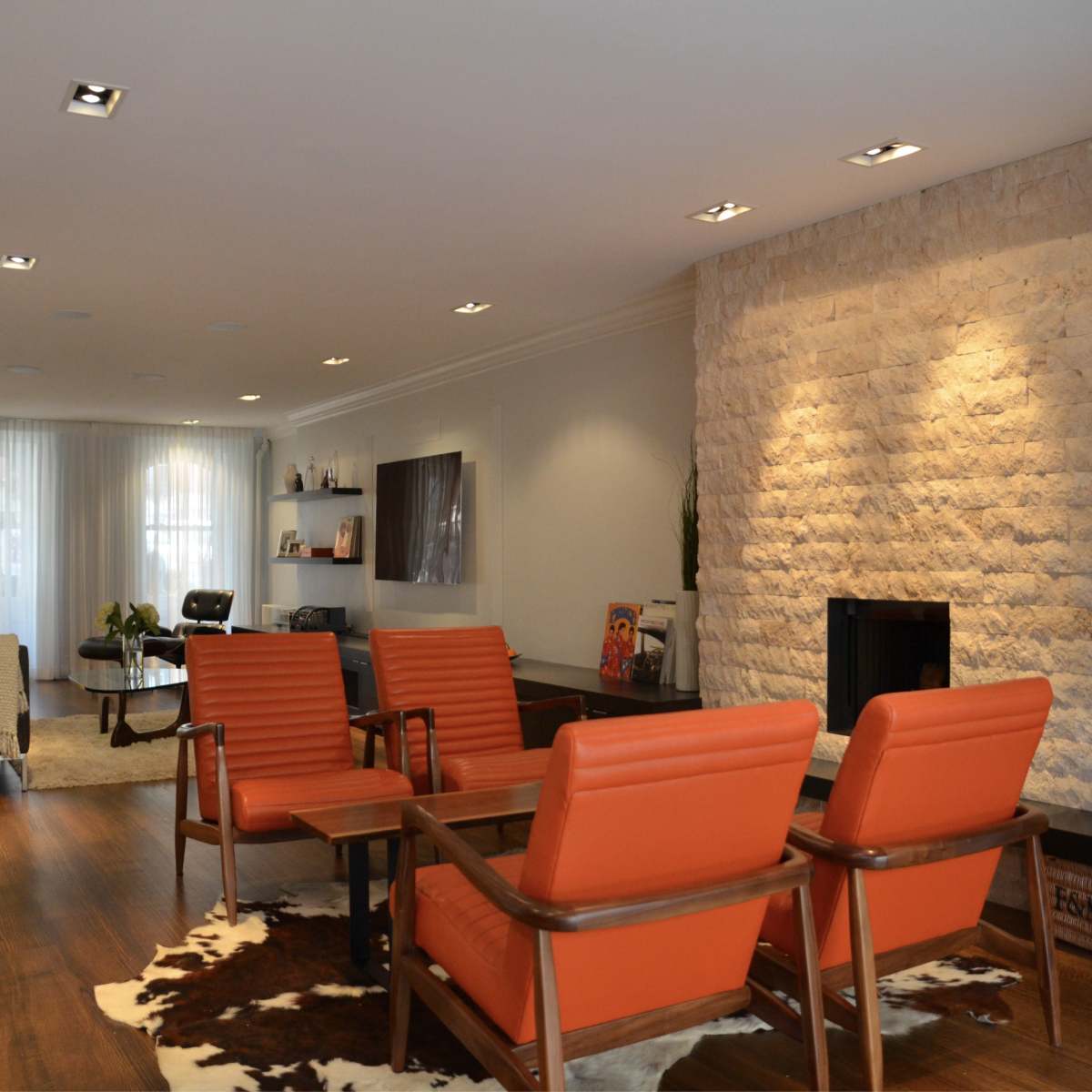 A modern living room features orange armchairs arranged around a coffee table, with a stone fireplace and a light carpeted area in the background.