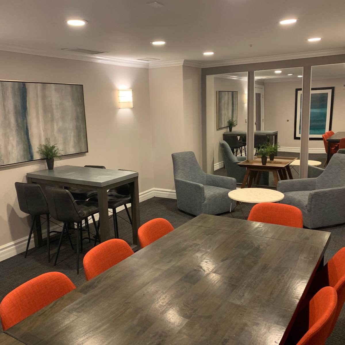 A modern lounge area featuring two seating arrangements, a large wooden table surrounded by orange chairs, and two gray armchairs near a small round table.