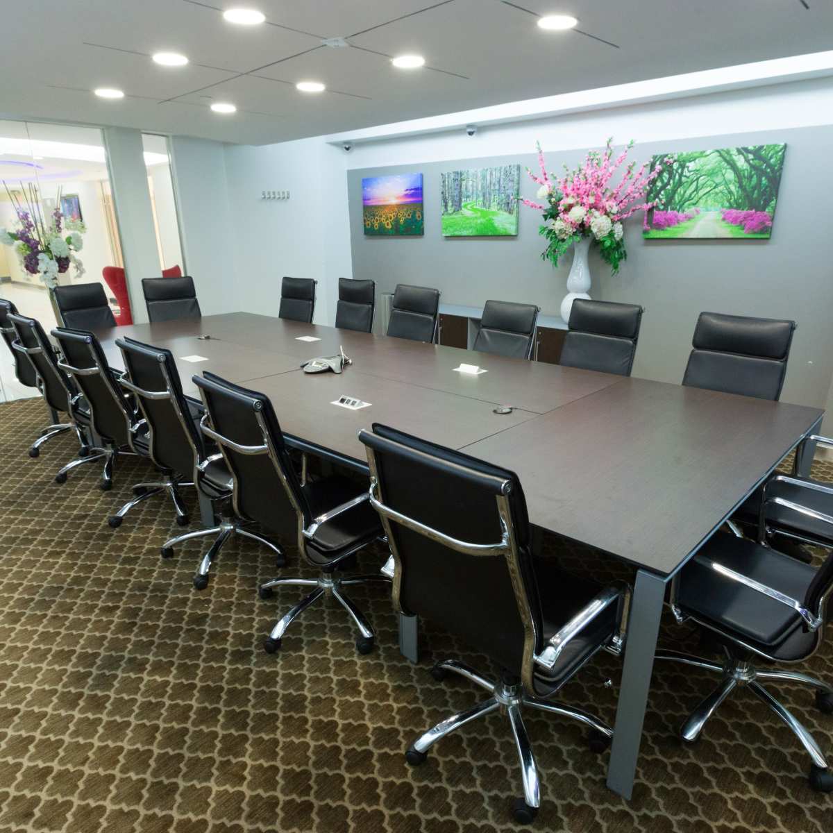 A long conference table with black chairs is set up in a modern meeting room, adorned with floral artwork on the walls.