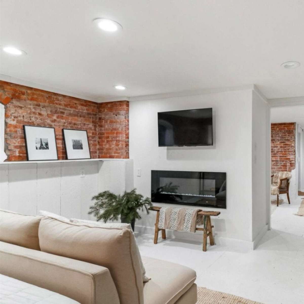 A modern basement area featuring exposed brick walls, a mounted television, a linear electric fireplace, and neutral-colored furnishings.
