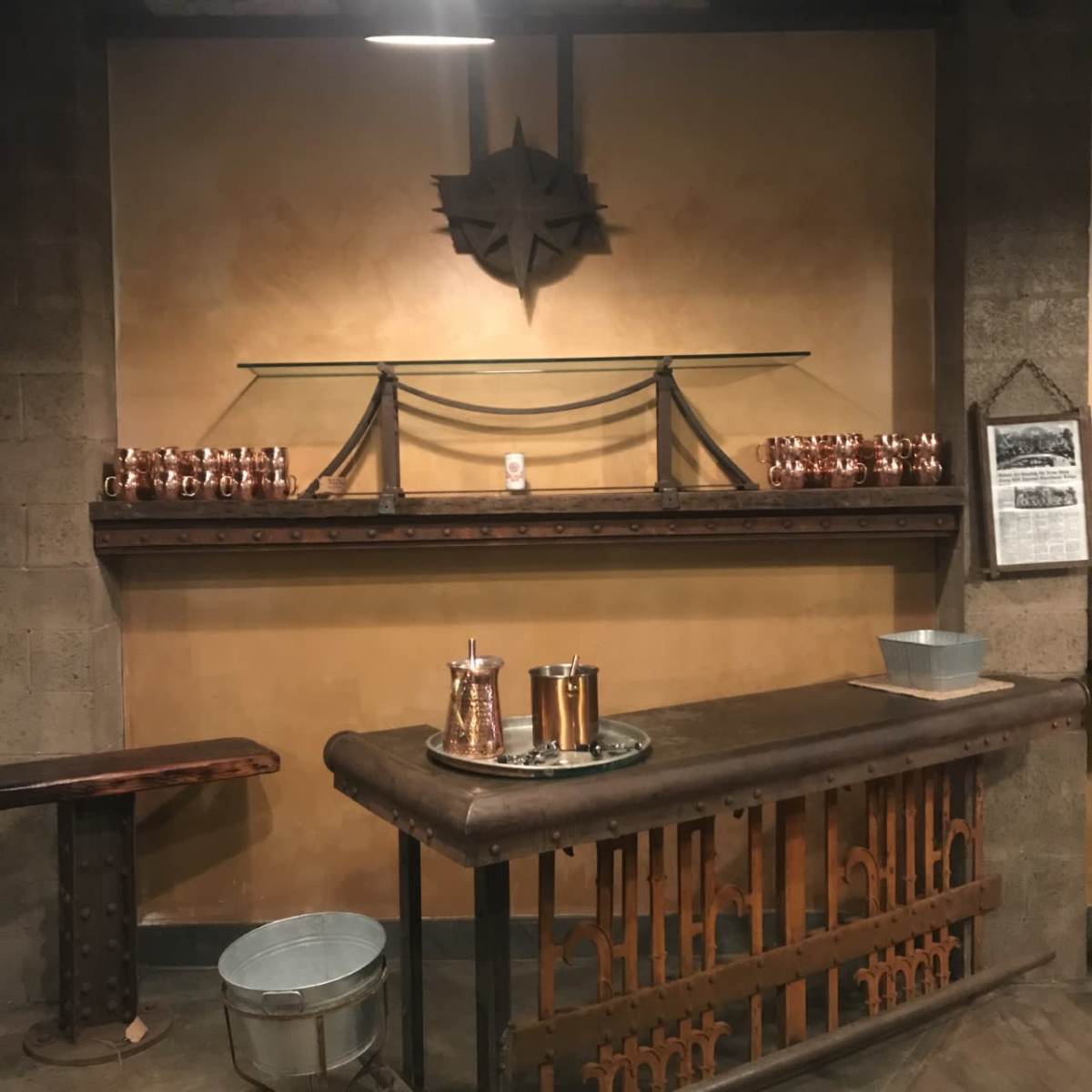 A rustic bar area with a wooden counter, metal accents, and a shelf displaying copper mugs and beverage containers against a textured wall.