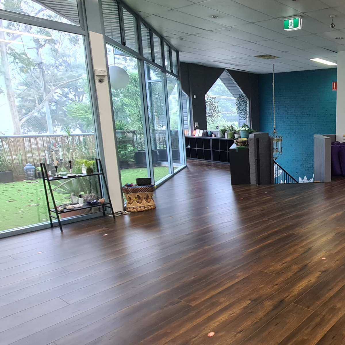 A spacious, empty room with wooden flooring, large windows revealing greenery outside, and minimal furnishings along one wall.