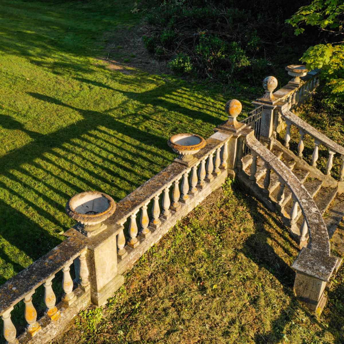 A stone balustrade with curved sections and ornamental pots, casting long shadows on a grassy lawn.