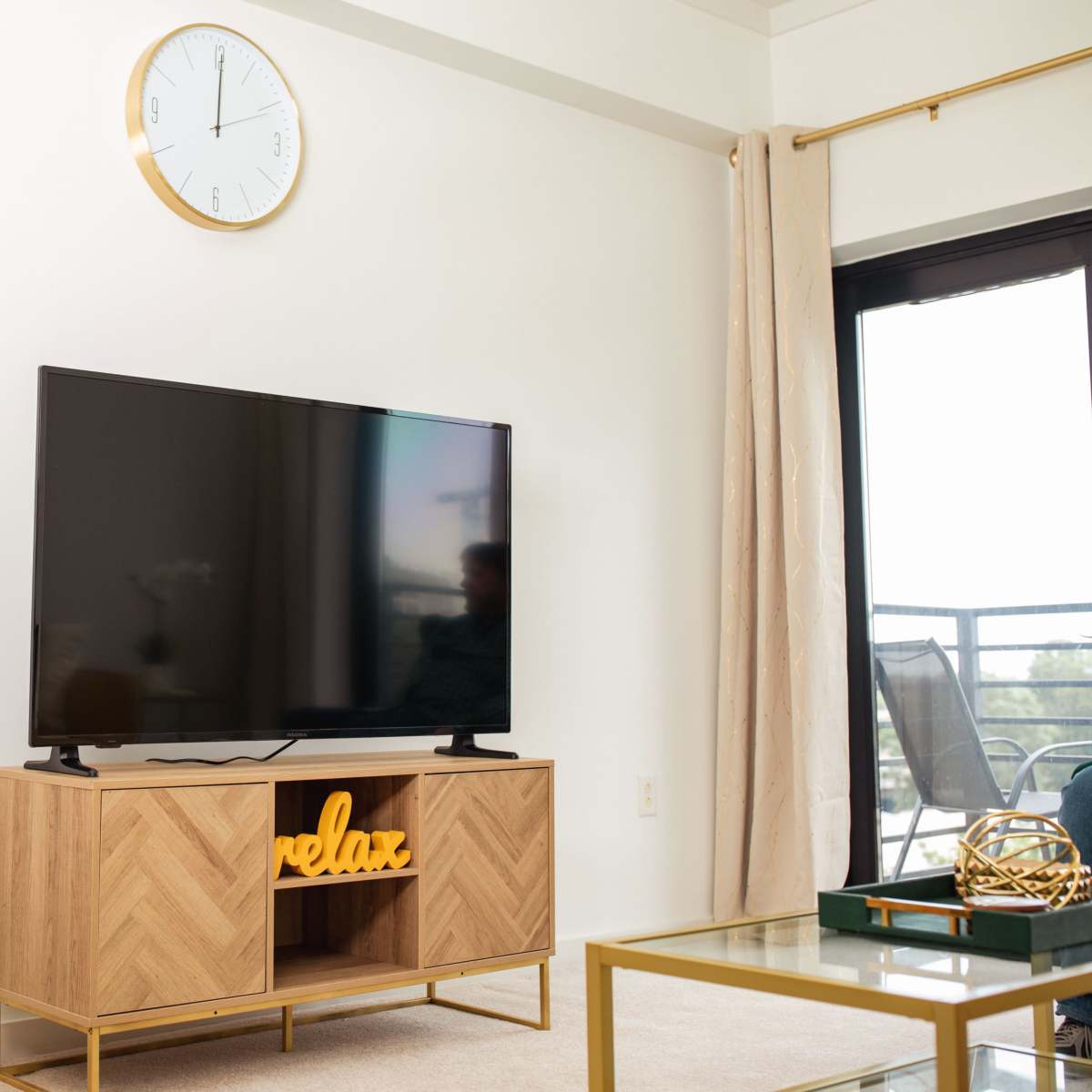 A modern living room features a television on a wooden stand with the word "relax" displayed, a wall clock, and a large window with a balcony view.
