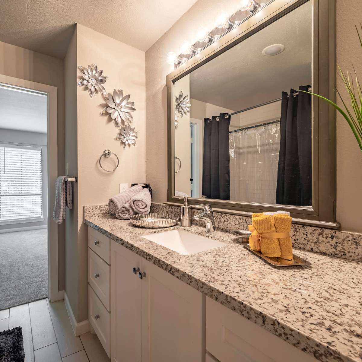 A modern bathroom features a double vanity with a mirrored wall and decorative elements, leading to an adjoining room visible through an open door.