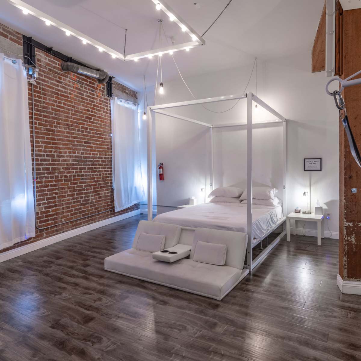 A minimalist bedroom featuring a four-poster bed, wooden floors, a brick wall, and soft lighting.