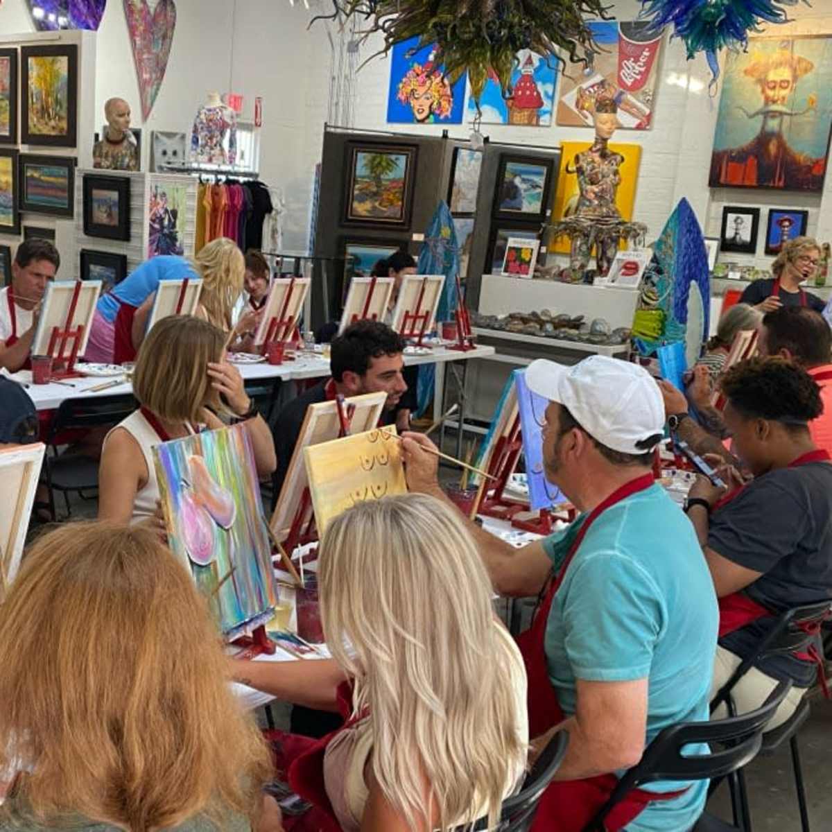 A group of people sits at tables in an art studio, each focused on painting on canvases while surrounded by various artworks displayed on the walls.