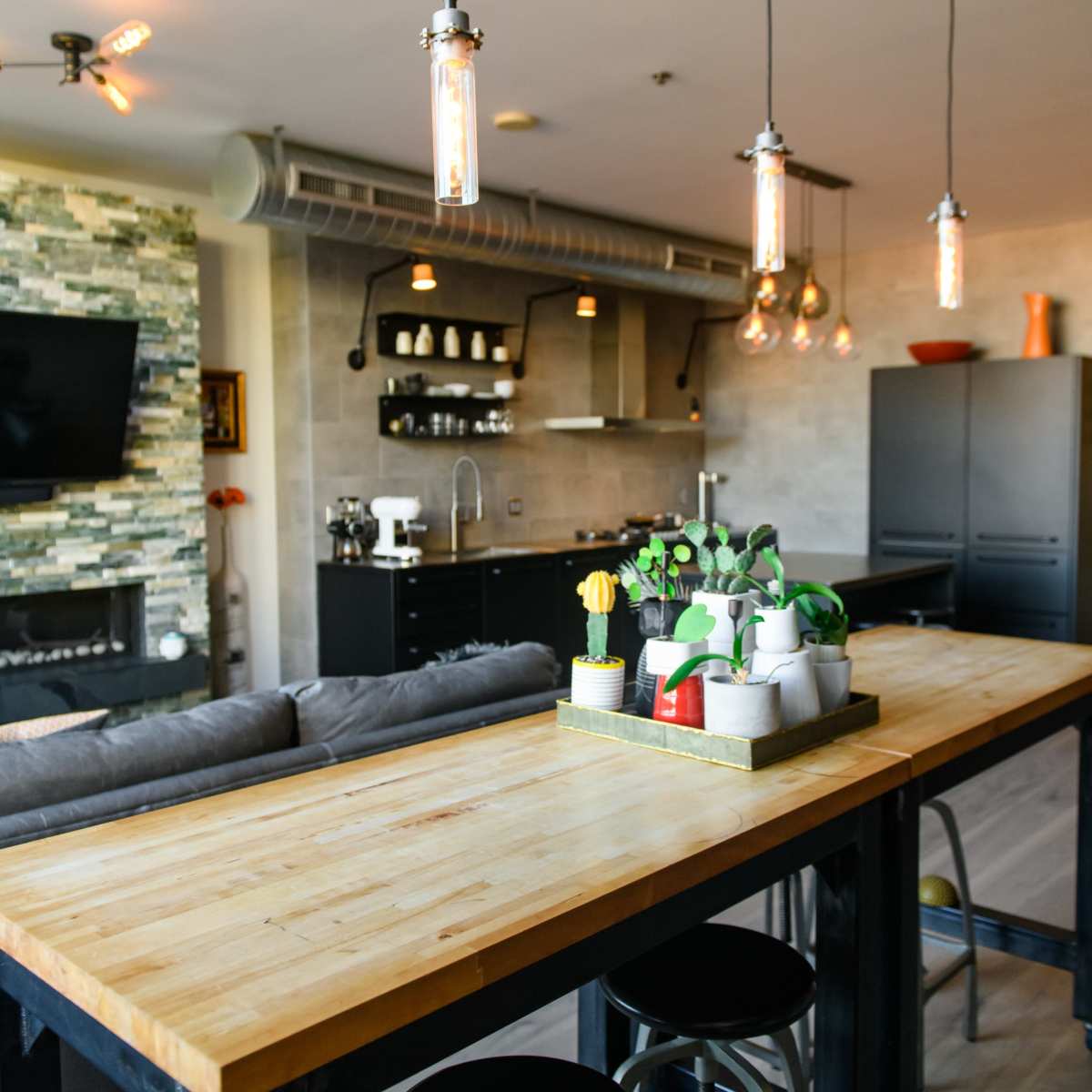 A modern kitchen features a wooden dining table with metal stools, pendant lighting, and a stone accent wall.