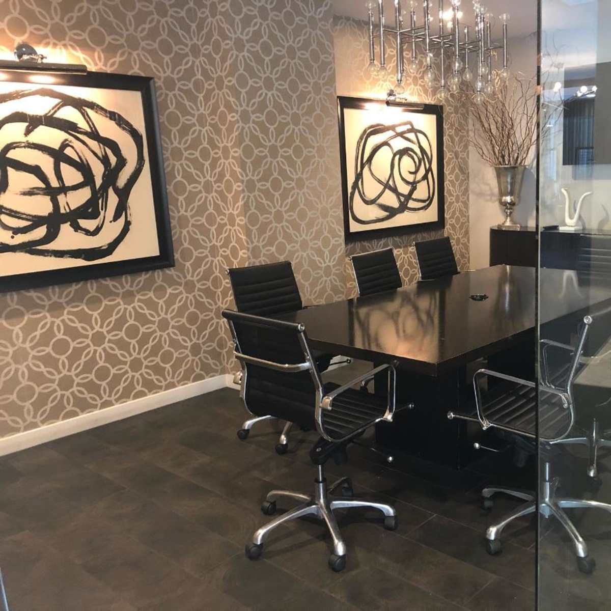 A modern conference room features a large black table surrounded by silver office chairs, with abstract wall art and a large screen on one side.