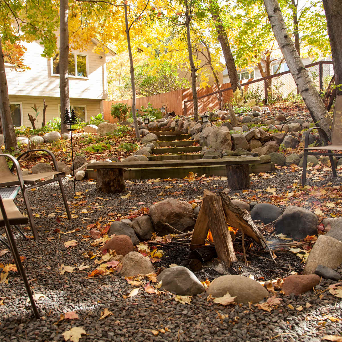A circular fire pit surrounded by wooden logs and stones is set in a backyard garden adorned with autumn leaves and stone pathways.