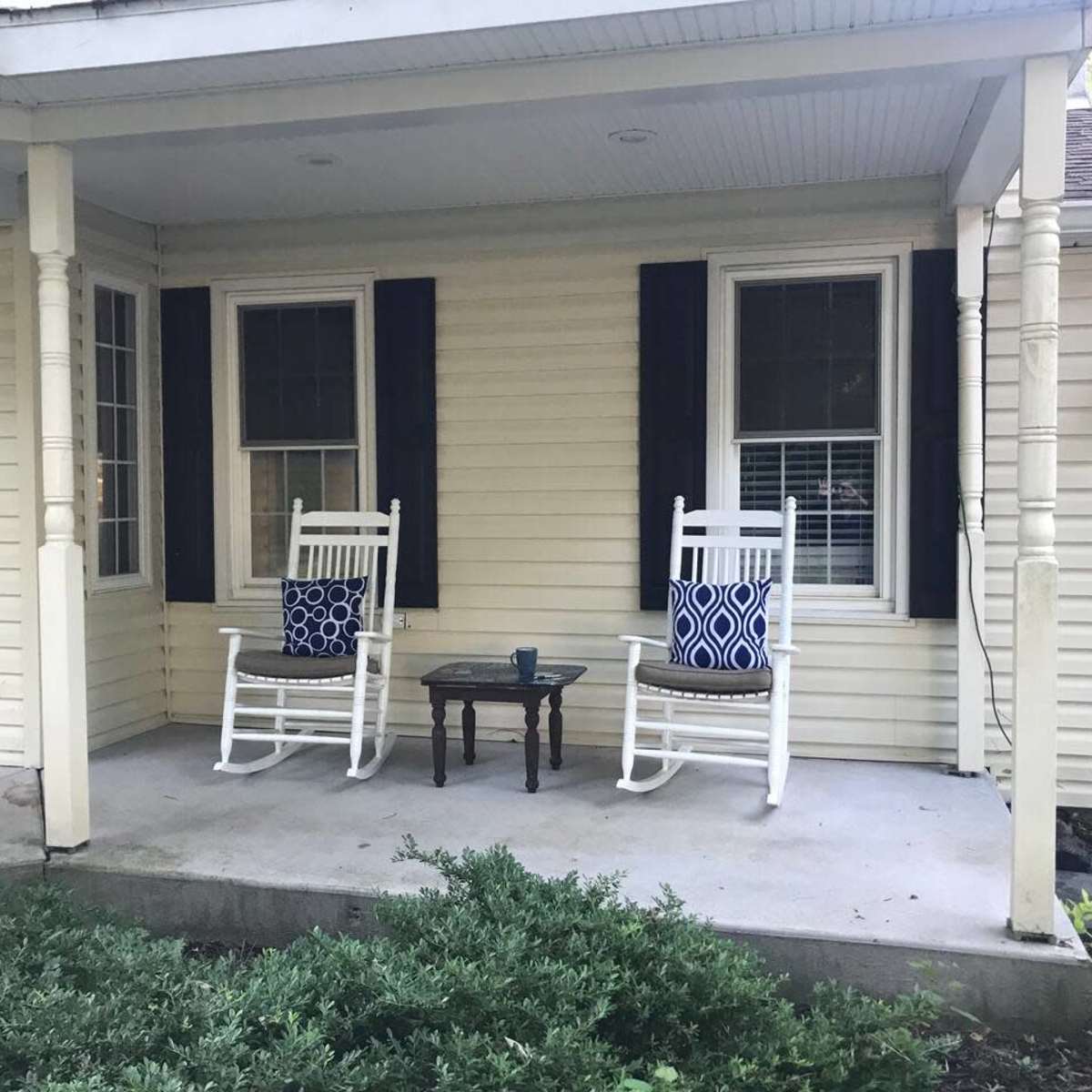 A front porch with two white rocking chairs, each adorned with blue patterned cushions, flanking a small wooden table.