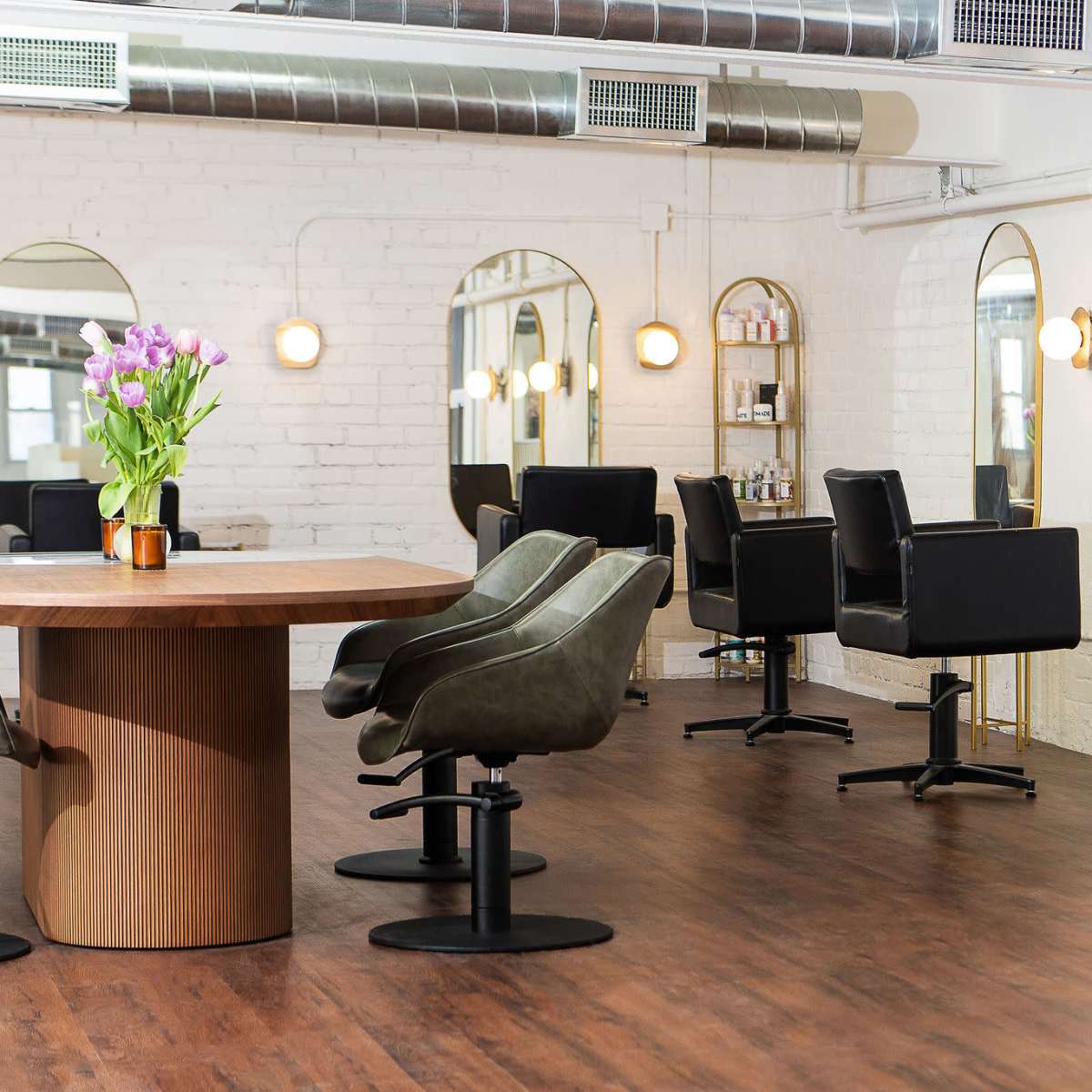 A modern salon featuring a central round table with a vase of flowers, several styling chairs, and large mirrors along a white brick wall.