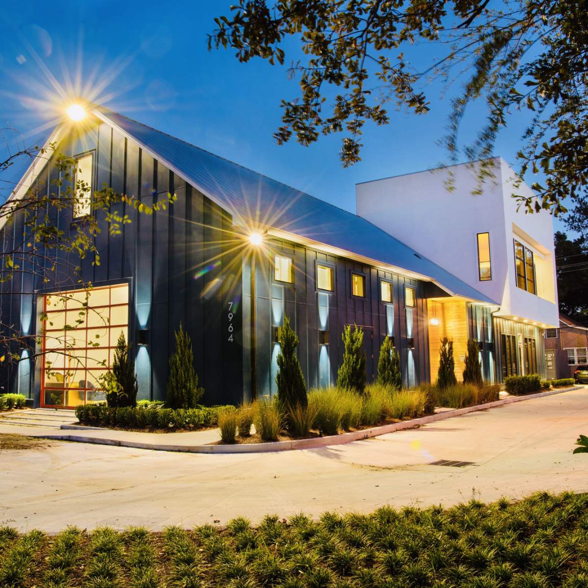A modern building with a black exterior, large windows, and a white section, illuminated at dusk with landscaping and a driveway in the foreground.