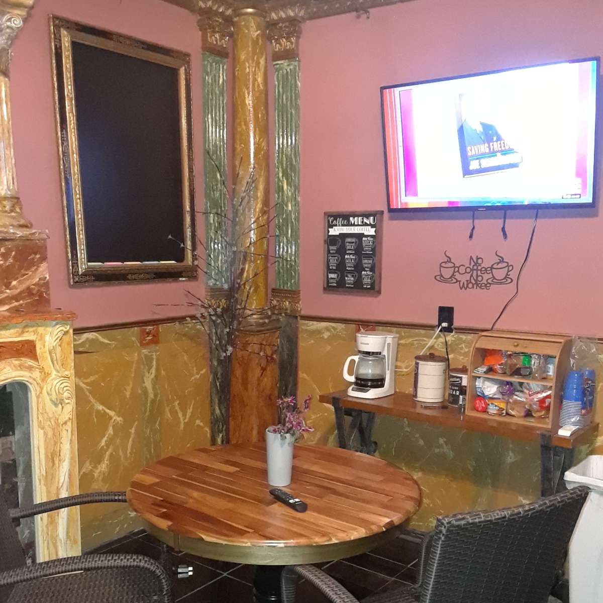 A small seating area with a wooden table, two wicker chairs, a coffee maker, snacks, and a TV mounted on the wall.