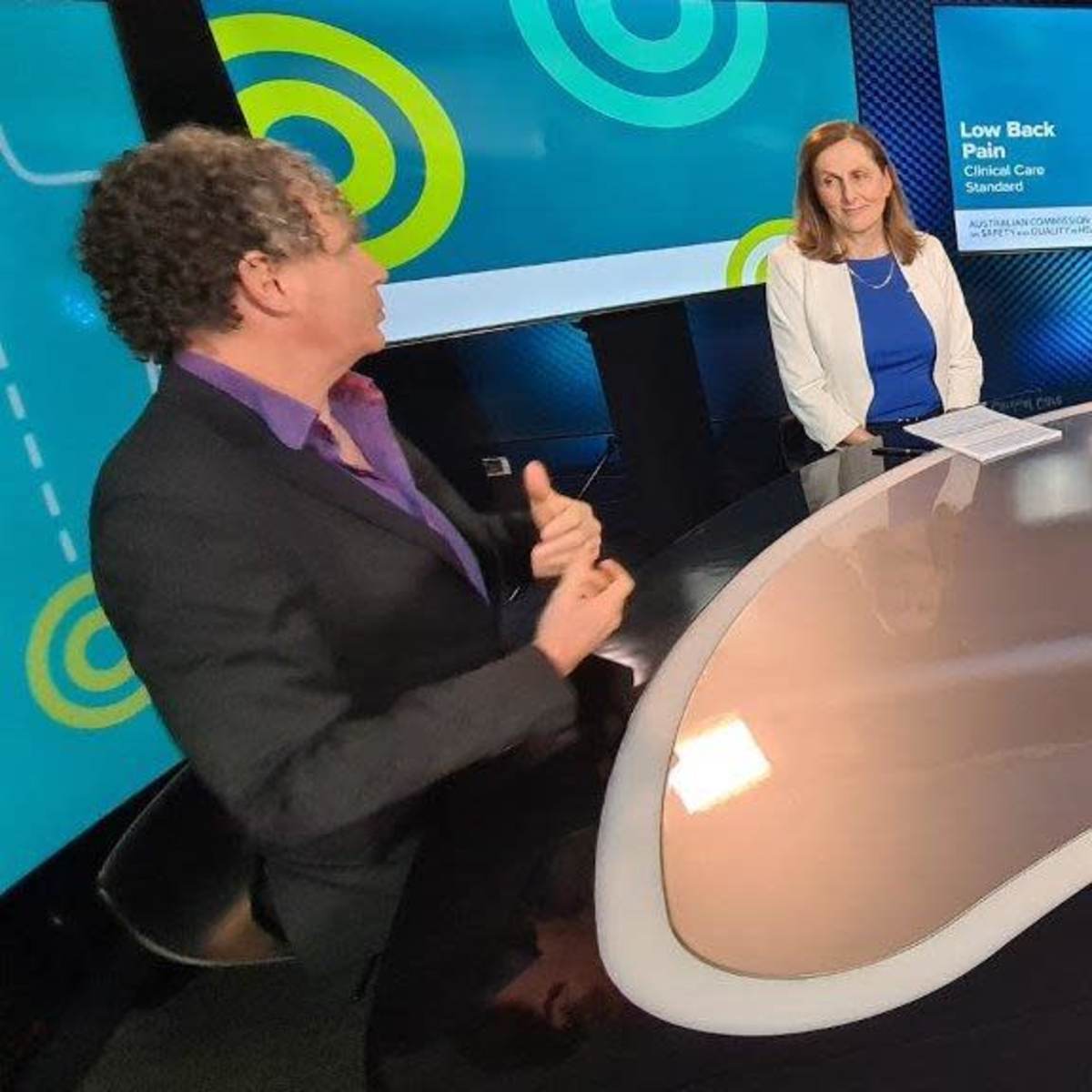 A group of five people is seated around a modern, curved table in a studio setting, discussing a topic related to a "Lawn Back Plan" presentation, with graphics in the background.