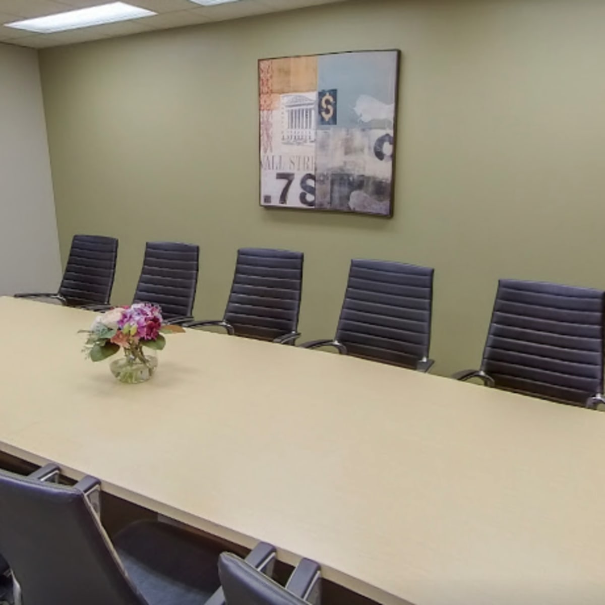 A conference room features a long table surrounded by eight black chairs, with a floral centerpiece and a whiteboard on one wall.