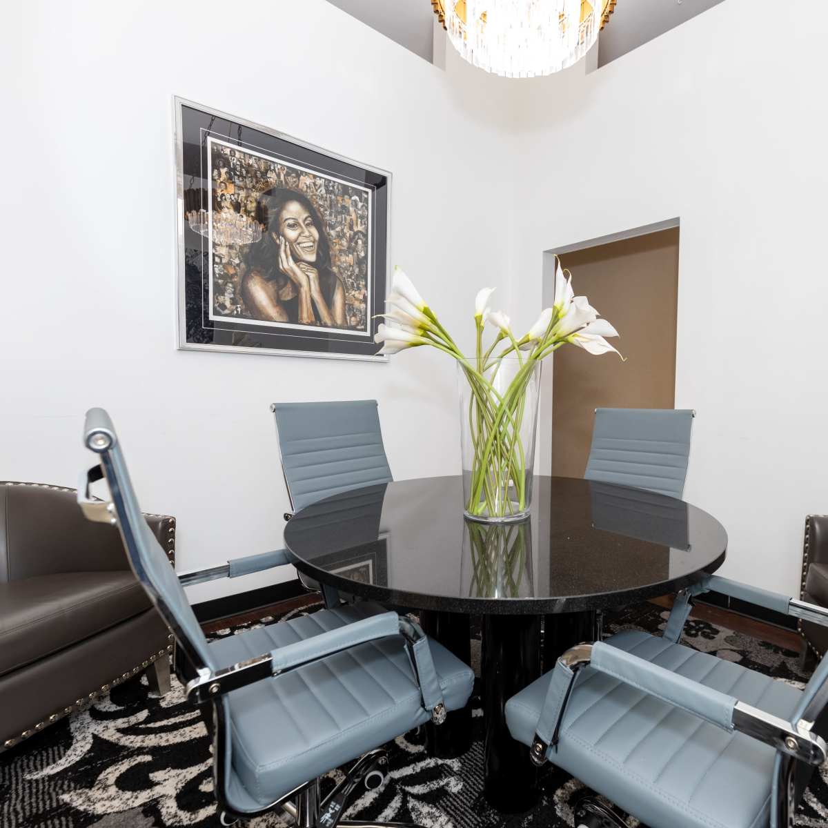 A modern conference room with a round black table surrounded by four blue and gray chairs and a large vase of white flowers on the table.