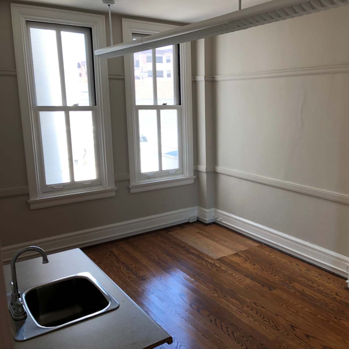 An empty room with wooden flooring, two windows, and a small kitchenette in one corner.