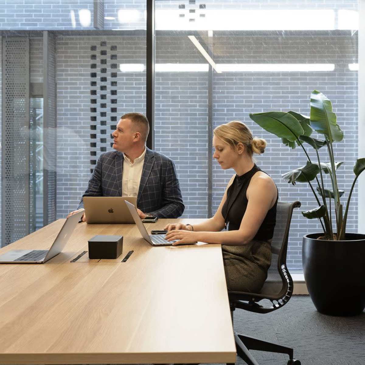A man writes on a whiteboard while two colleagues sit at a conference table, one working on a laptop and the other observing.