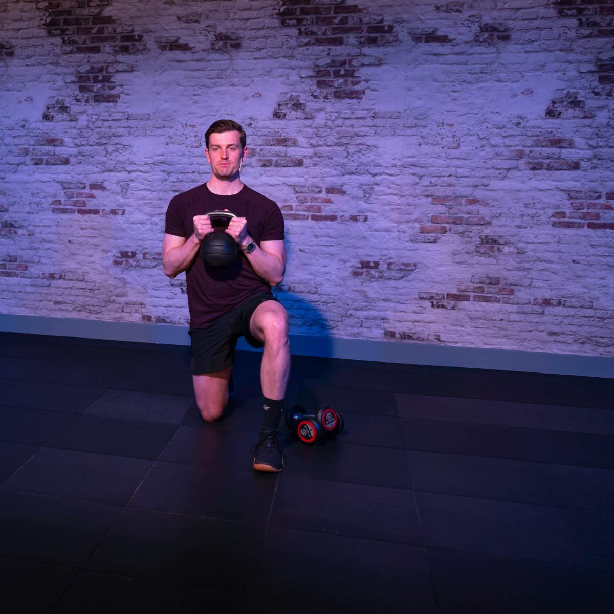A man is kneeling on one leg, holding a kettlebell, while being filmed by a camera on a tripod in a studio with a brick wall.