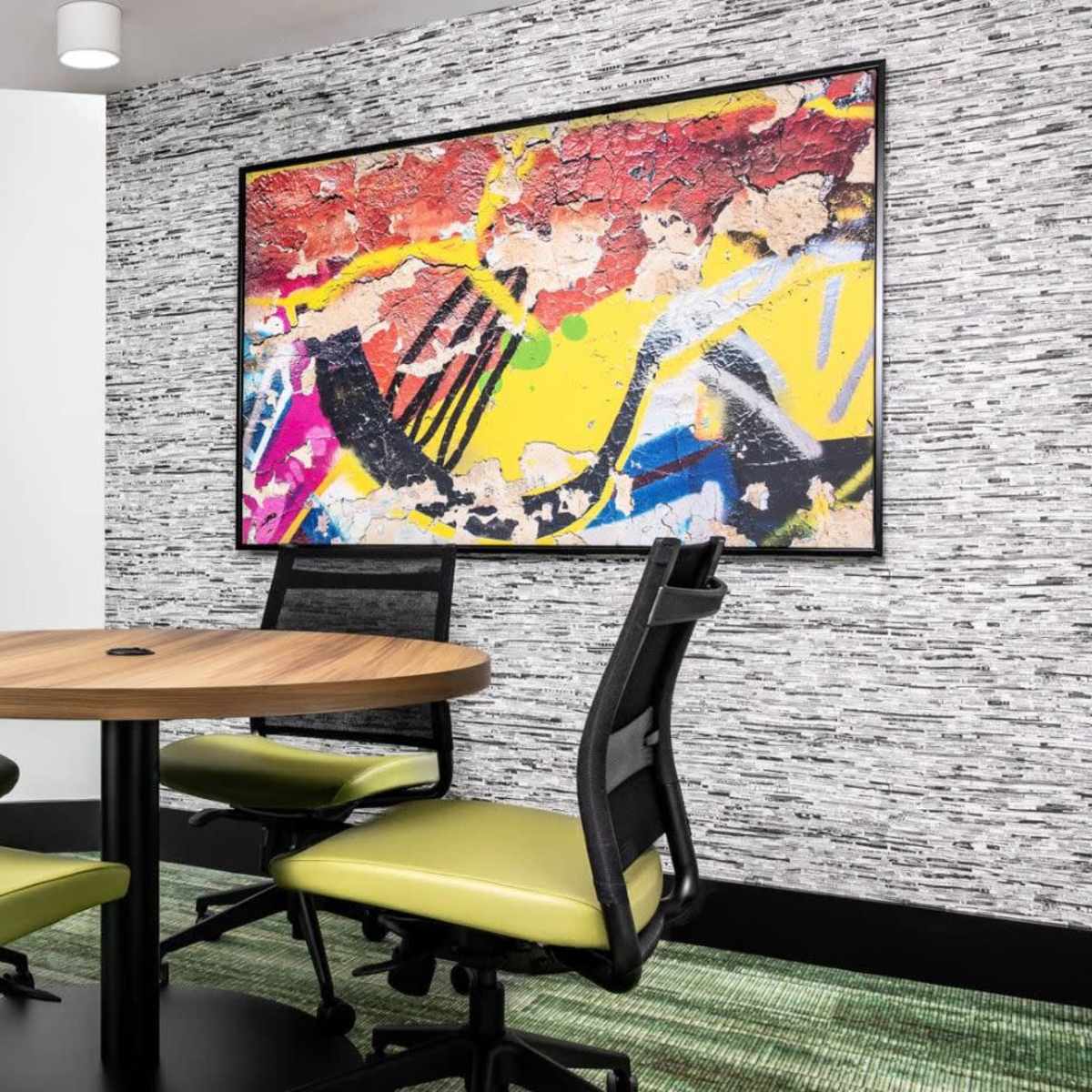 A conference room features a round wooden table surrounded by four black and green chairs, with a colorful abstract painting on the wall and textured wallpaper.