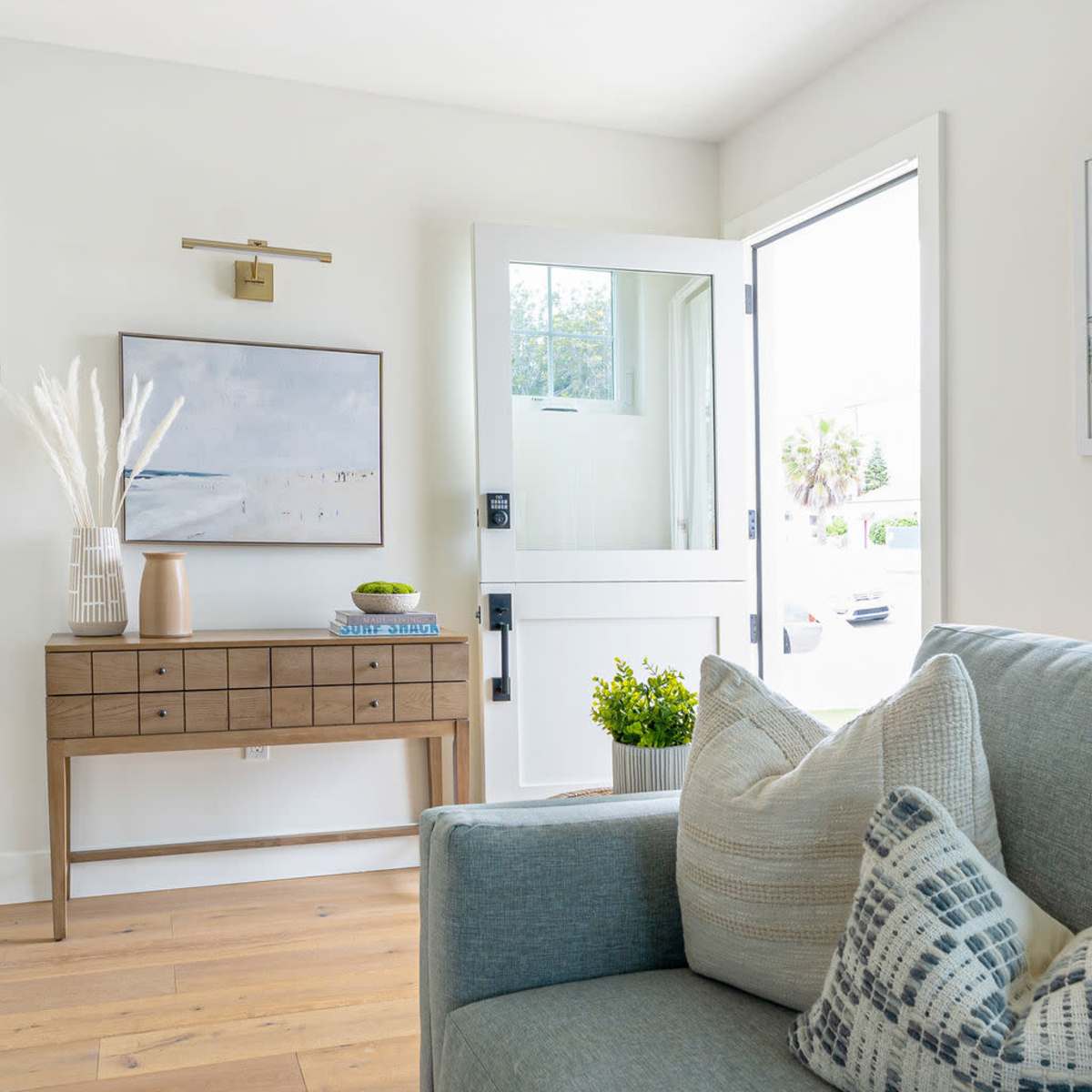 A bright living room features a light-colored sofa with decorative pillows, a wooden console table against the wall, and an open door leading to the outside.