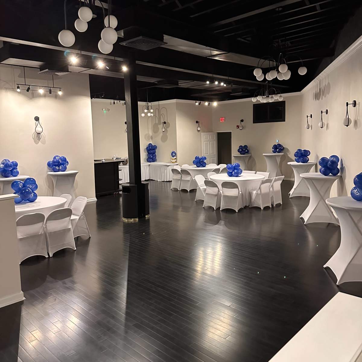 A spacious event venue with white tables adorned with blue balloons, set against a black wooden floor and illuminated by ceiling lights.