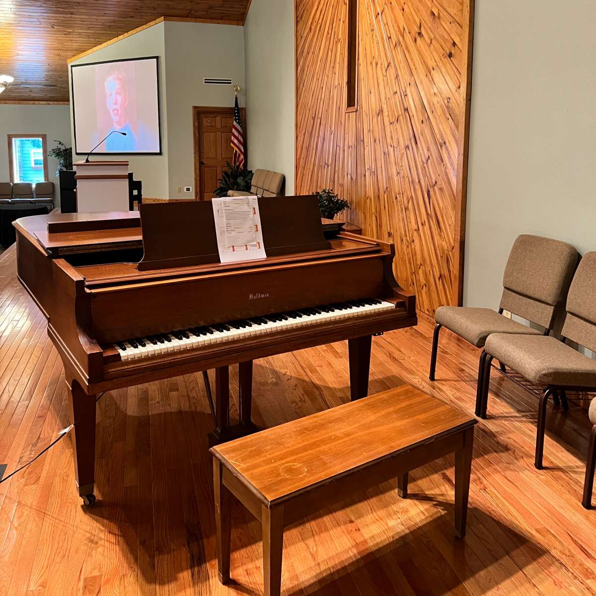 A wooden grand piano is placed in front of a backdrop featuring a large cross and a projection screen in a church setting.
